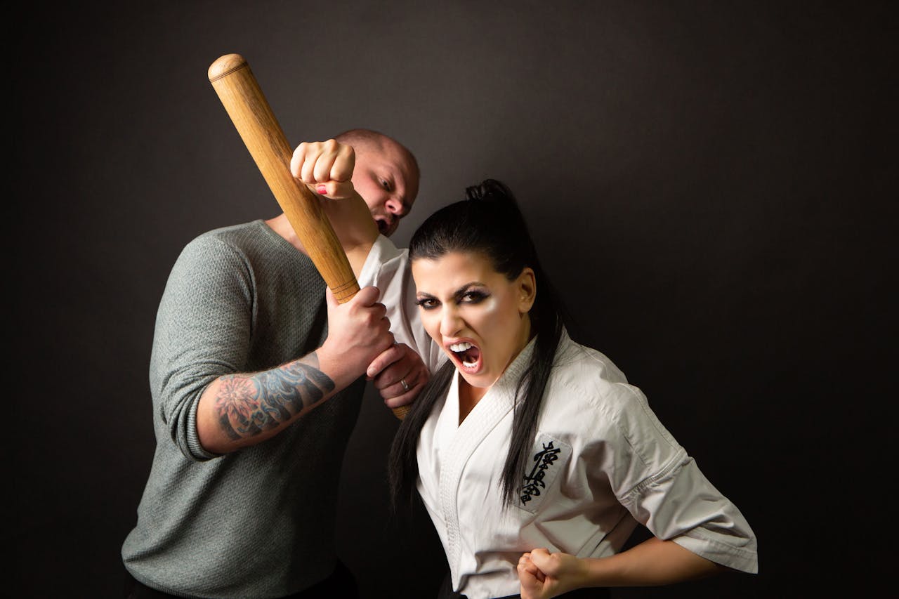 Woman in karate uniform demonstrates self-defense gracefully against a tattooed man with bat.